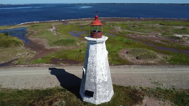 Approche a&eacute;rienne en drone du vieux phare Chiasson-Office d&eacute;peintur&eacute; par le temps en bardeaux de c&egrave;dre blancs et rouges au pied d'un chemin non-pav&eacute; longeant des milieux humides et le bord de la mer. 