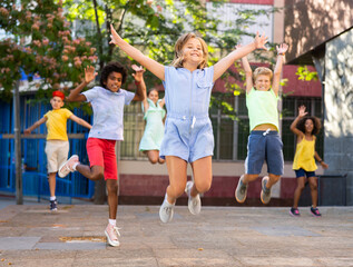 Fototapeta premium Cheerful emotional preteen girl gaily spending time with friends on summer day, jumping together on city street..
