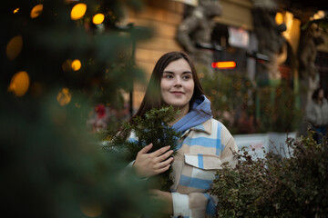 Street city photo. Young adult childfree brunette woman with green fir branch near Christmas tree....
