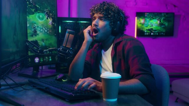Indian Arabian Latino American man headphones playing video game online computer pc cyberspace yawning tired bored drinking coffee male streamer guy display keyboard monitor technology gamer networks