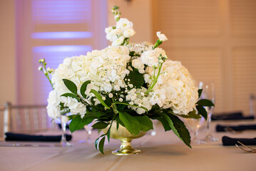 White roses and green leaf floral centerpiece in gold vase on table with bokeh background purple