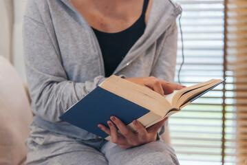 Obraz premium Happy asian woman use headphones listen to music reading book sitting on cozy sofa in living room at home. Female asian people work at home research book read magazine smiling happy reader lifestyle