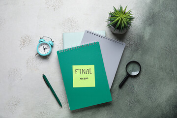 Sticky note with text FINAL EXAM, notebooks, magnifier and alarm clock on grunge background