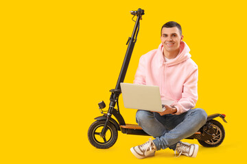 Young man with electric scooter using laptop on yellow background