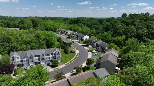 Suburban neighborhood nestled in lush greenery in Pennsylvania. Rows of townhouses line a winding street. Verdant trees surround the residential area. Aerial.