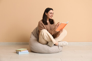 Young woman sitting on pouf and reading book near beige wall
