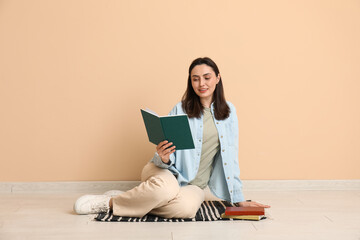 Young woman sitting on floor and reading book near beige wall