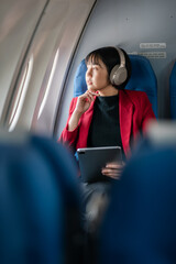 A businesswoman in a red blazer and headphones holding a tablet, gazing thoughtfully out of an airplane window during a flight.