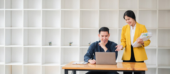 Two business professionals working together in a modern office setting, discussing work over a laptop and documents.