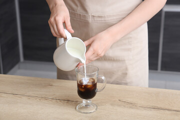 Young woman pouring milk from jug into glass with ice coffee in kitchen