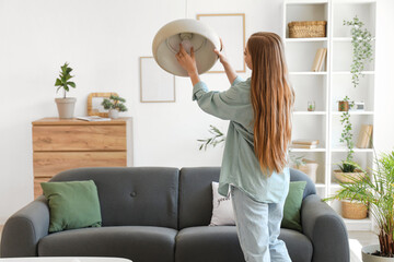 Young woman screwing light bulb into ceiling lamp at home, back view
