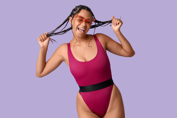Obraz premium Beautiful young African-American woman in stylish purple swimsuit and sunglasses on lilac background