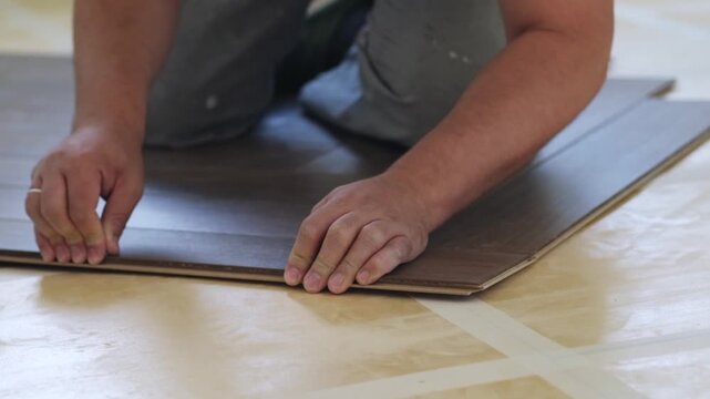 Close-up of a man kneeling and installing laminate flooring in a home. Focus on hands carefully placing panels. Perfect for DIY, renovation, and interior design projects