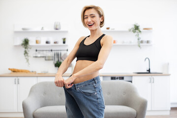 Woman smiling while showing loose jeans indicating weight loss. Celebrating fitness success in bright home interior. Fit woman wearing sports bra standing in living room with plants and shelves.