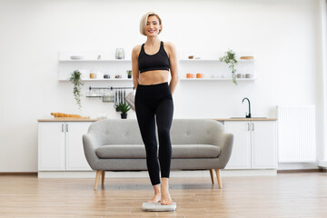 Fit woman in sportswear standing on scale in bright living room measuring weight. Concept of...