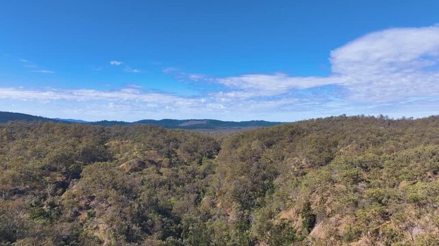 Aerial rises above the Barimoon area, and forest covered hills to expose the Boyd Burnett Rail Trail cutting through the woodlands with views to Kalpower State Forest Central Queensland Australia.