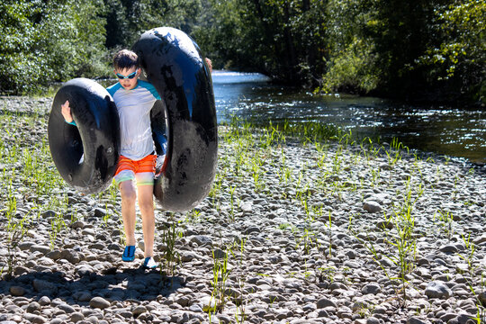 Boy carrying two large tubes after tubing, walking on a rocky riverbed