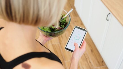 Woman in fitness clothing tracking daily calorie intake on smartphone while holding bowl of fresh salad. Concept of healthy eating, fitness, nutrition, and technology.
