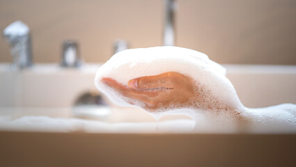 People hand is playing shower soap foam bubbles during take a shower in bathtub. Beauty, healthcare and skincare concept photo. Close-up and selective focus at the hand.