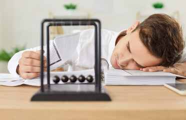 Tired businessman sitting at work desk with Newton's cradle pendulum at foreground, putting head on...