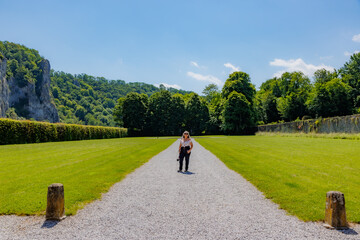 Fototapeta premium Gravel path with female tourist standing in garden outside Freyr Castle, lush trees and rocky mountain against blue sky in blurred background, sunny summer day in Namur, Wallonia, Belgium