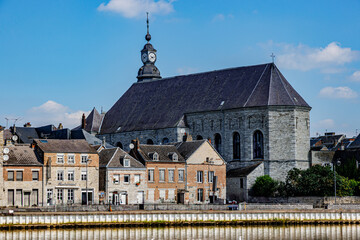 Naklejka premium Old buildings and back view of St. Hilarius church against blue sky in background, cityscape of Givet town, Meuse river in foreground, sunny summer day in Ardennes department, France