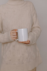 Woman in warm knitted sweater holds white mockup cup of coffee or tea