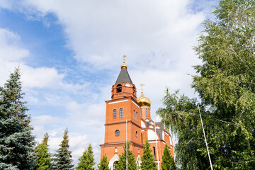 Obraz premium A large red brick church featuring an impressive bell tower is beautifully surrounded by lush green trees that enhance its stunning appearance and contribute to the serene atmosphere