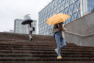Girl with orange umbrella walking down the steps on buildings background, people during rain in city