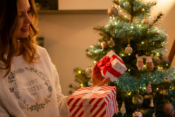 Chica en pijama navide&ntilde;o sujetando cajas de regalo al lado del &aacute;rbol de navidad. 