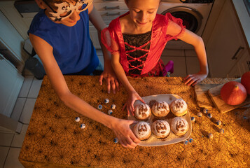 girl and brother trying buns with ghost images, they are dressed in Halloween costumes, enjoying holiday treats in decorated dark kitchen, warm cozy atmosphere, holiday mood, 