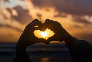 A heart shape made with hands, set against the backdrop of a beautiful sunset over the ocean.