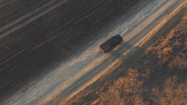 Aerial shot of pickup truck driving through rural path near ploughed field. Flying over off road vehicle moving on route near farmland. Black car moving along road near plowed meadow at autumn