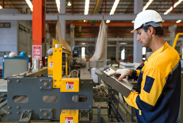 Young hispanic labor dressed in polyester jacket uniform, hardhat and protective glove checks the orderliness of the machine. Working atmosphere in metal sheet factory with large industrial machines.