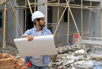 Young engineer in a construction helmet holding a floor plan while looking at the progress of real estate projects. The construction site has scaffolding made of wood and a pile of bricks.