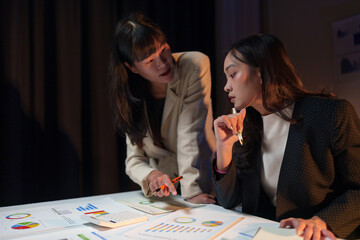 Two businesswomen working overtime, having discussions, talking, brainstorming, pointing at graphs...