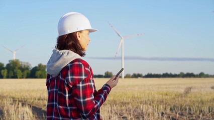 Woman engineer is using a smartphone in a field with wind turbines as the sun sets. Concept of clean energy and engineering audit