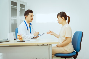 A male Asian pediatrician in a white lab coat sits at his desk, warmly greeting a middle-aged...