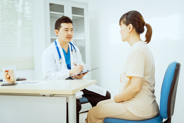 Obraz premium A male Asian pediatrician in a white lab coat sits at his desk, warmly greeting a middle-aged pregnant woman for a prenatal consultation, offering care and support in a clinical setting.