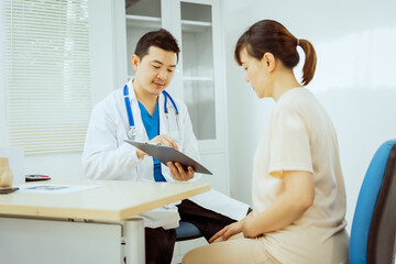 Fototapeta premium A male Asian pediatrician in a white lab coat sits at his desk, warmly greeting a middle-aged pregnant woman for a prenatal consultation, offering care and support in a clinical setting.