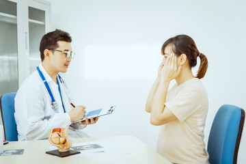 A male Asian pediatrician in a white lab coat sits at his desk, warmly greeting a middle-aged pregnant woman for a prenatal consultation, offering care and support in a clinical setting.