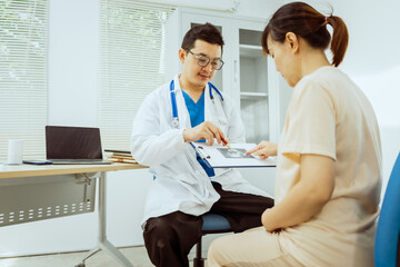 Obraz premium A male Asian pediatrician in a white lab coat sits at his desk, warmly greeting a middle-aged pregnant woman for a prenatal consultation, offering care and support in a clinical setting.
