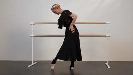 A beautiful ballerina in a black dress dances in a white sunny room with a black floor next to a ballet barre.