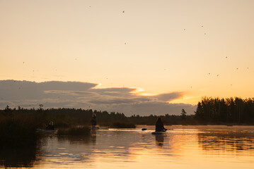 A serene scene of people on SUP boards watching the sunrise over a calm lake, surrounded by dense forest. The sky is painted with soft hues, reflecting on the water.