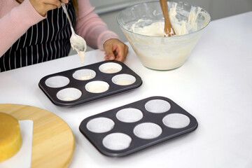 A person is pouring batter into a cupcake tray, preparing to bake. A sponge cake sits nearby.