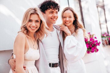 Young three stylish friends posing in the street. Fashion man and two cute female dressed in casual summer clothes. Smiling models having fun. Cheerful women and guy outdoors, in sunny day, near wall