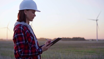 Female engineer taking notes on a clipboard on a field with wind turbines, as the sun sets. Concept of clean energy and engineering audit
