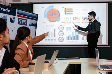 A cheerful and confident Asian businessman stands, presenting bar charts data from the projector...