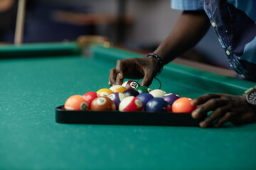 Close-up of hands arranging colorful billiard balls in triangular rack on green pool table in dimly...
