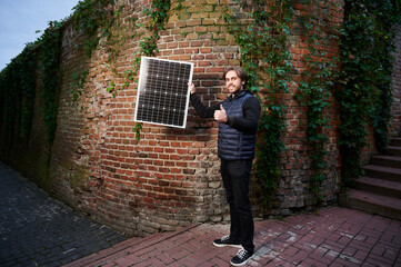 Man holding photovoltaic solar panel in front of old brick wall covered in green ivy, giving thumbs...